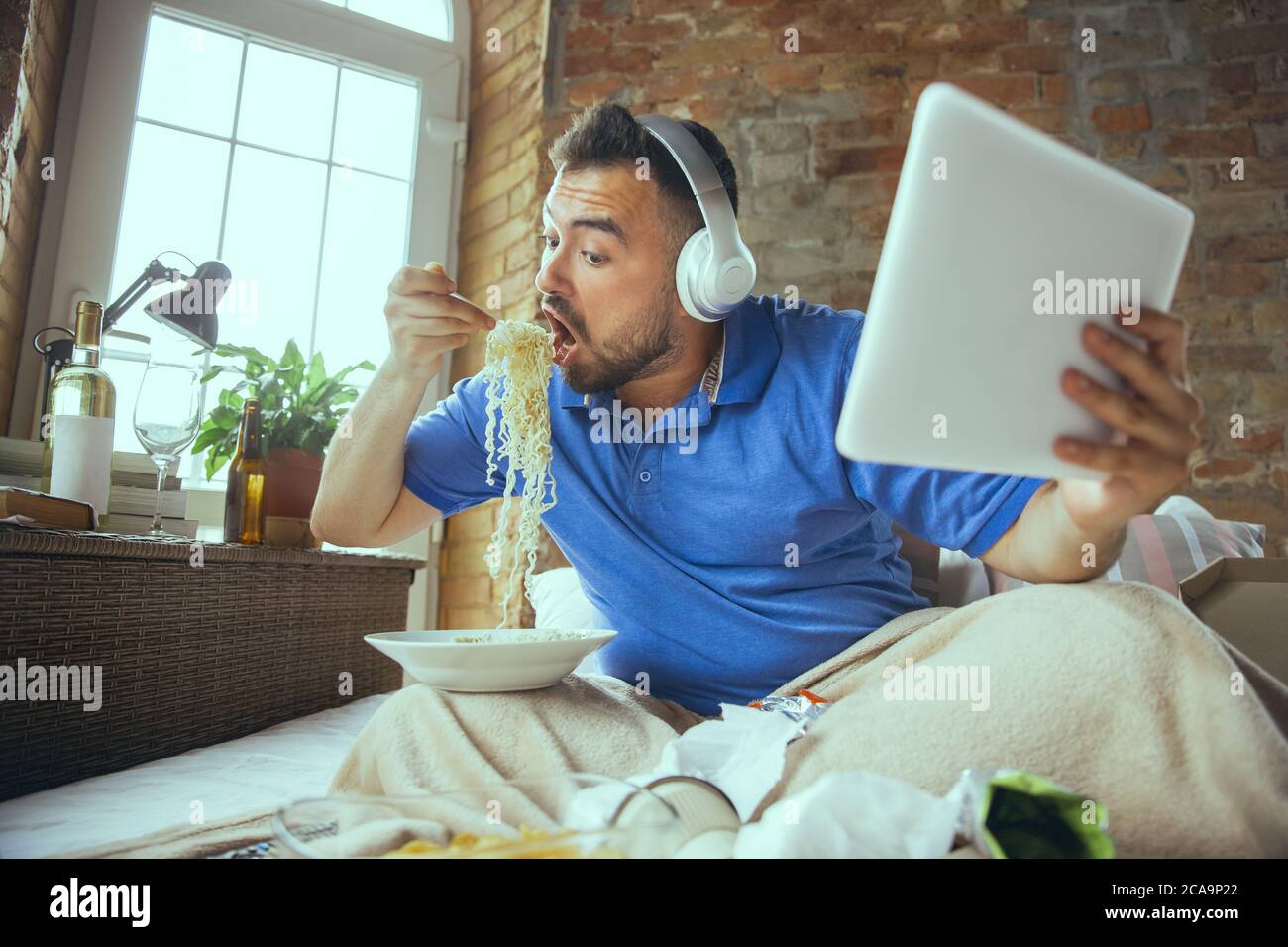 Eating instant noodle, watching match, championship, cinema. Lazy man living in his bed surrounded with messy. No need to go out to be happy. Using gadgets, watching movie and series, looks emotional. Stock Photo