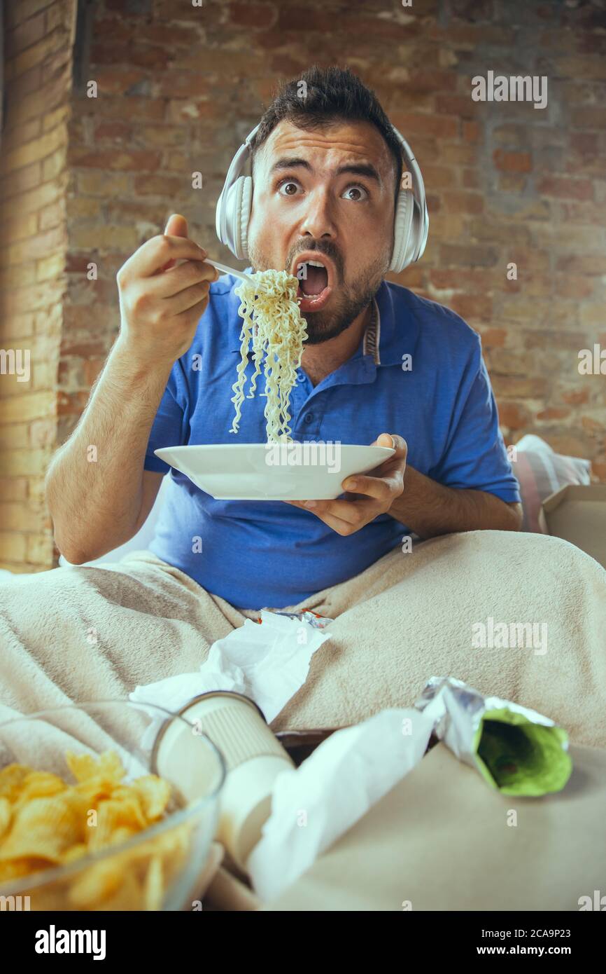 Shocked eating instant noodle, watching TV. Lazy man living in his bed surrounded with messy. No need to go out to be happy. Using gadgets, watching movie and series, looks emotional. Fast food. Stock Photo