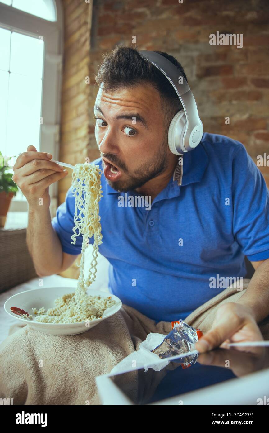 Eating instant noodle, watching match, championship, cinema. Lazy man living in his bed surrounded with messy. No need to go out to be happy. Using gadgets, watching movie and series, looks emotional. Stock Photo