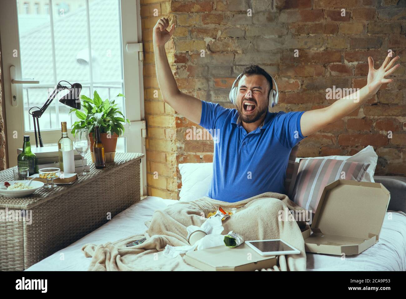 Crazy happy, celebrating win of favourite team. Lazy man living in his bed surrounded with messy. No need to go out to be happy. Using gadgets, watching movie and series, looks emotional. Stock Photo