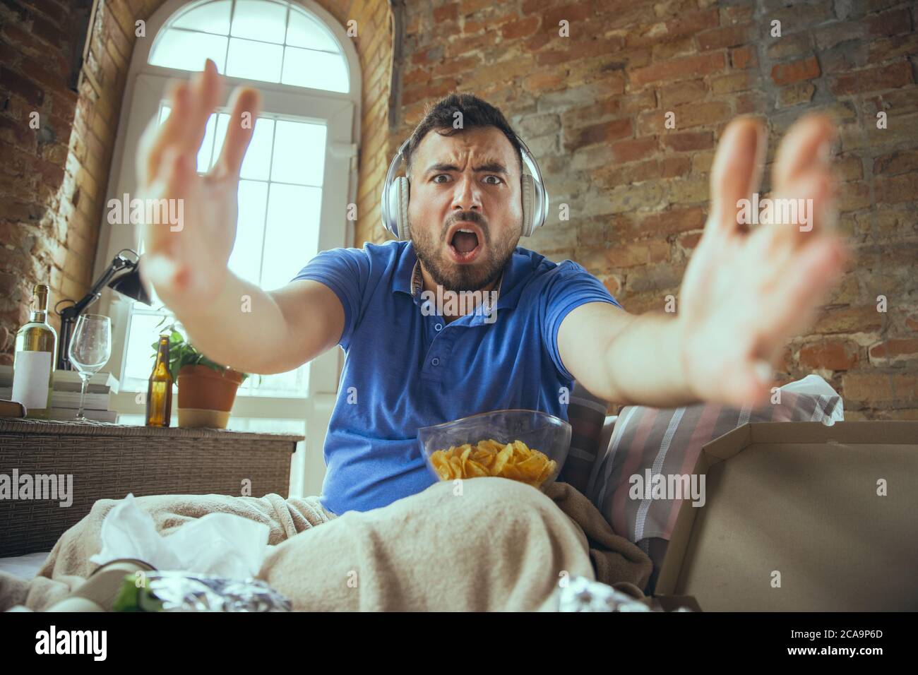 Excited watching match of favourite team. Lazy man living in his bed surrounded with messy. No need to go out to be happy. Using gadgets, watching movie and series, looks emotional. Fast food. Stock Photo