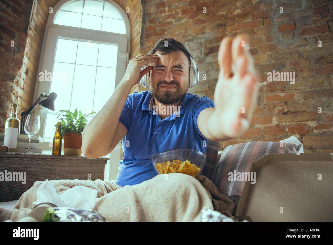 Disappointed watching match of favourite team. Lazy man living in his bed surrounded with messy. No need to go out to be happy. Using gadgets, watching movie and series, looks emotional. Fast food. Stock Photo