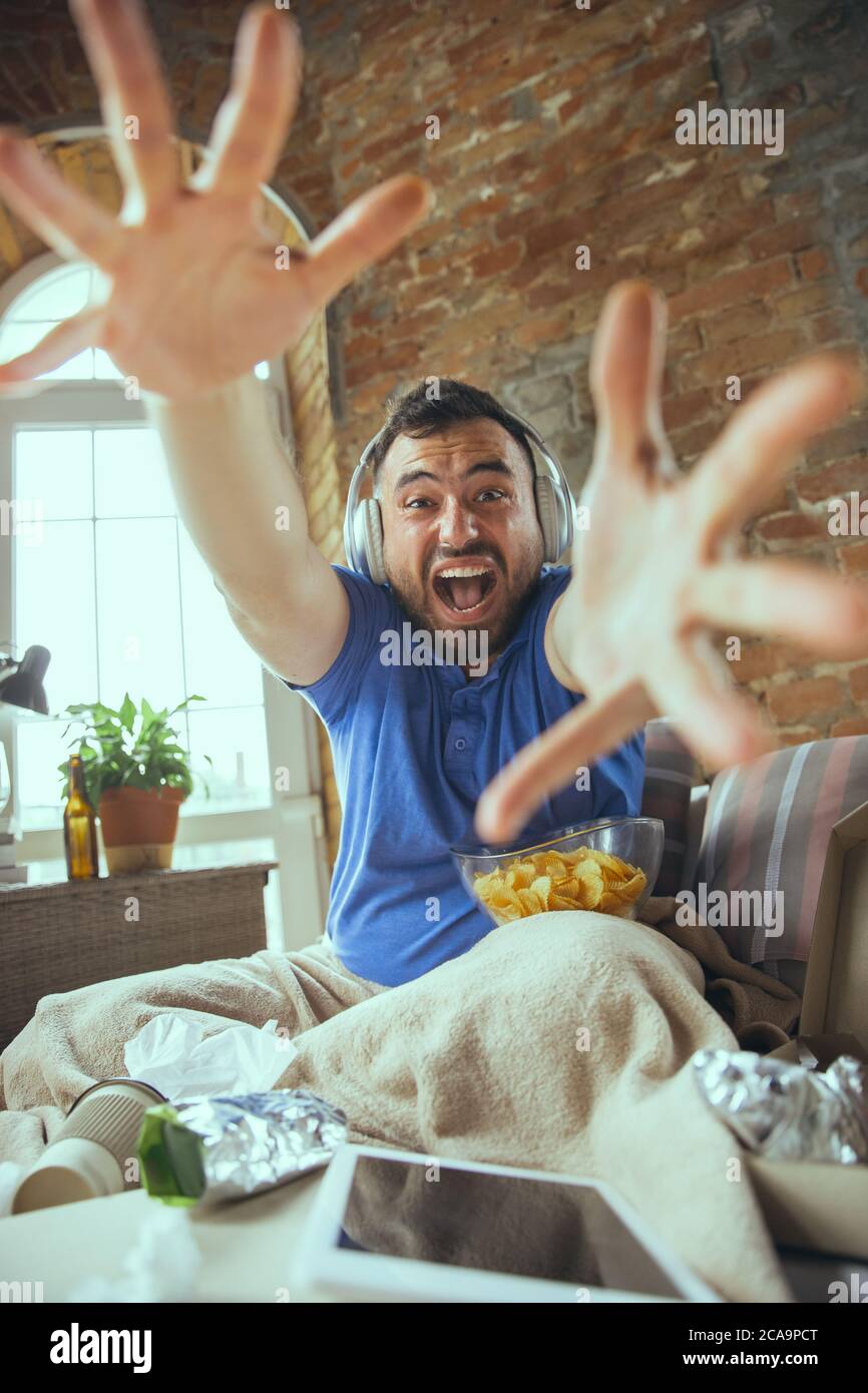 Crazy happy of favourite team. Videocall, social media. Lazy man living in his bed surrounded with messy. No need to go out to be happy. Using gadgets, watching movie and series, emotional. Fast food. Stock Photo