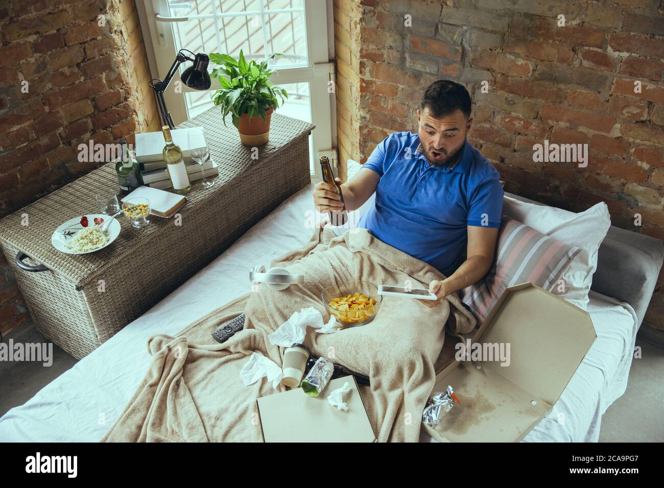 Excited cheering of favourite sport team, drinks beer. Lazy man living in his bed surrounded with messy. No need to go out to be happy. Using gadgets, watching movie and series, emotional. Fast food. Stock Photo