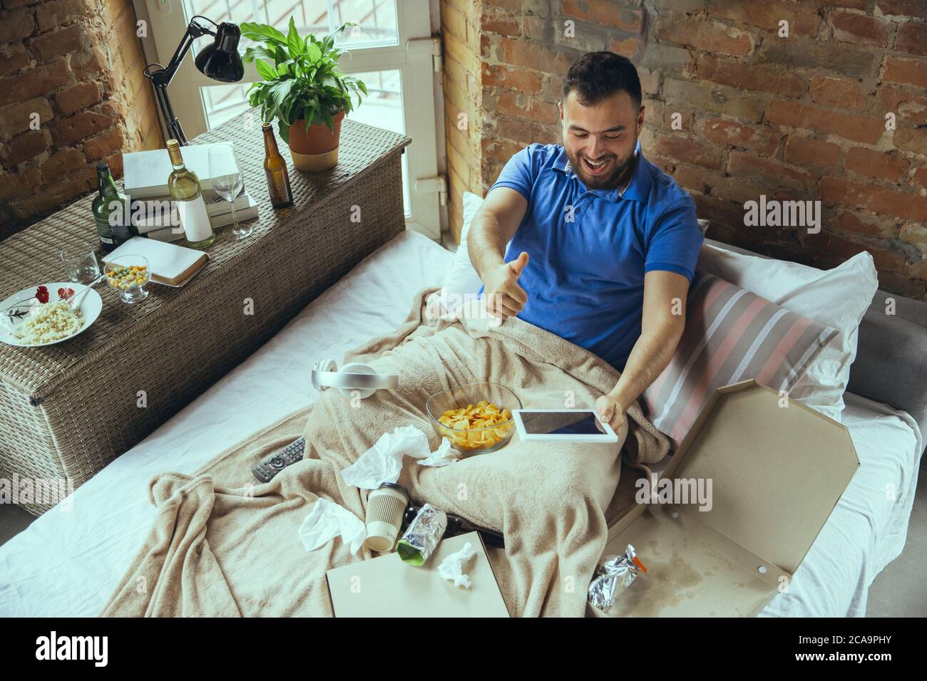 Excited cheering of sport team, thumb up. Lazy man living in his bed surrounded with messy. No need to go out to be happy. Using gadgets, watching movie and series, emotional. Fast food. Stock Photo