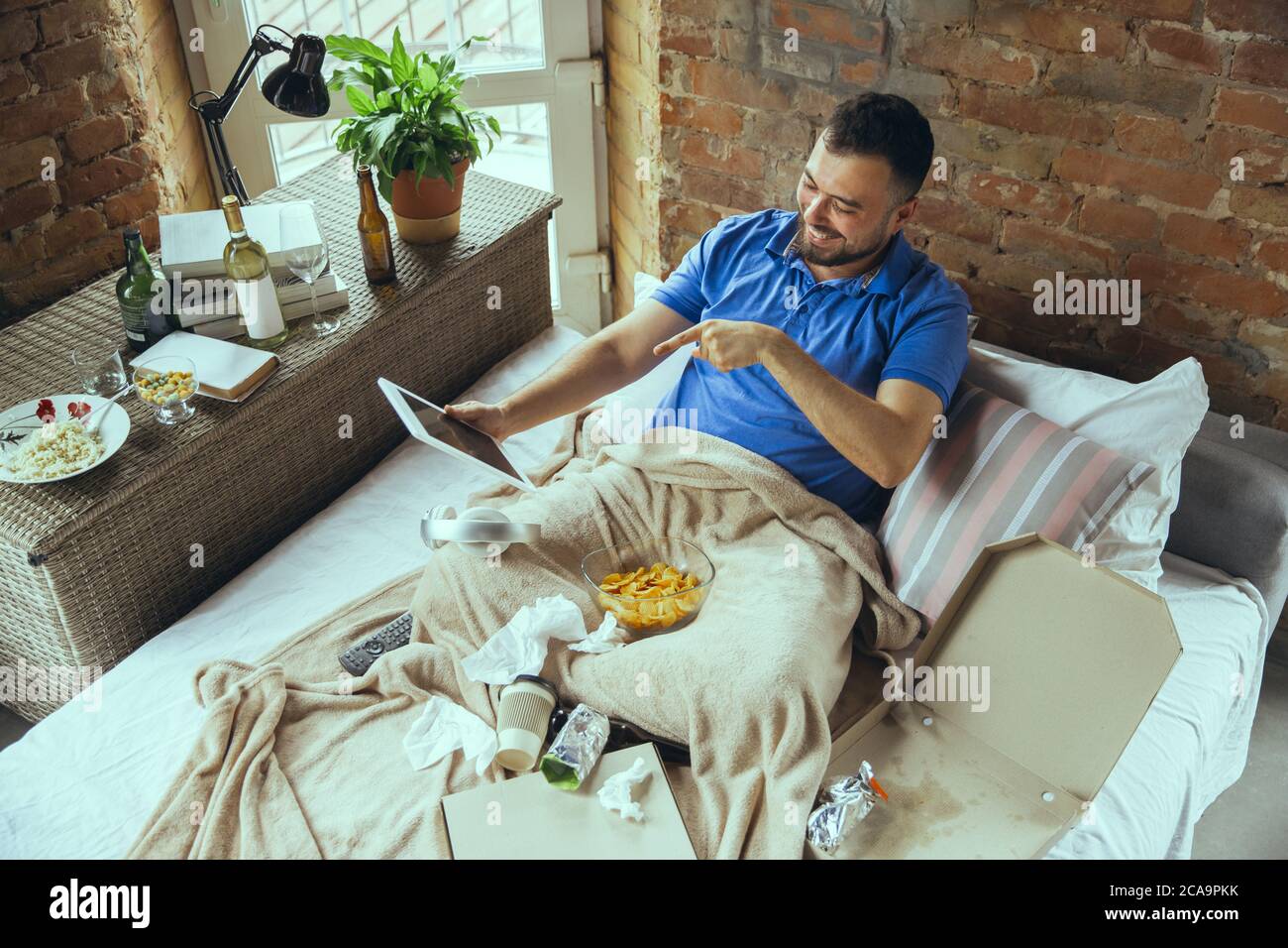 Smiling, pointing during videocall using tablet. Lazy man living in his bed surrounded with messy. No need to go out to be happy. Using gadgets, watching movie and series, emotional. Fast food. Stock Photo