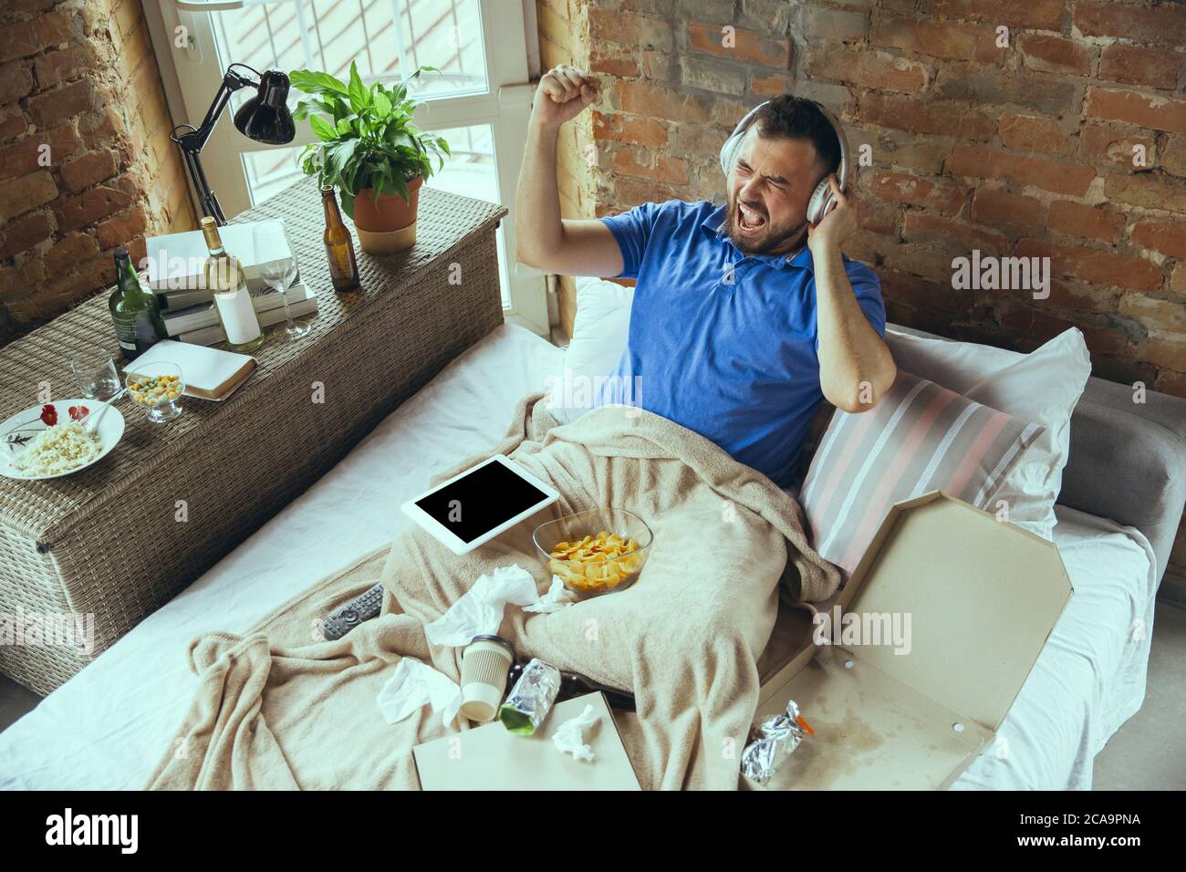 Crazy drive, listening to music and singing. Lazy man living in his bed surrounded with messy. No need to go out to be happy. Using gadgets, watching movie and series, emotional. Fast food. Stock Photo