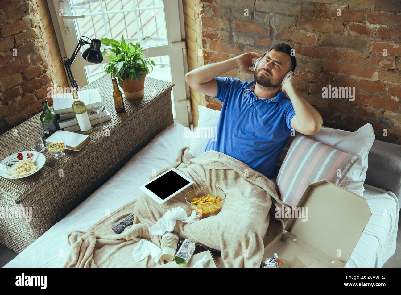 Joyful, inspired listening to music with headphones. Lazy man living in his bed surrounded with messy. No need to go out to be happy. Using gadgets, watching movie and series, emotional. Fast food. Stock Photo