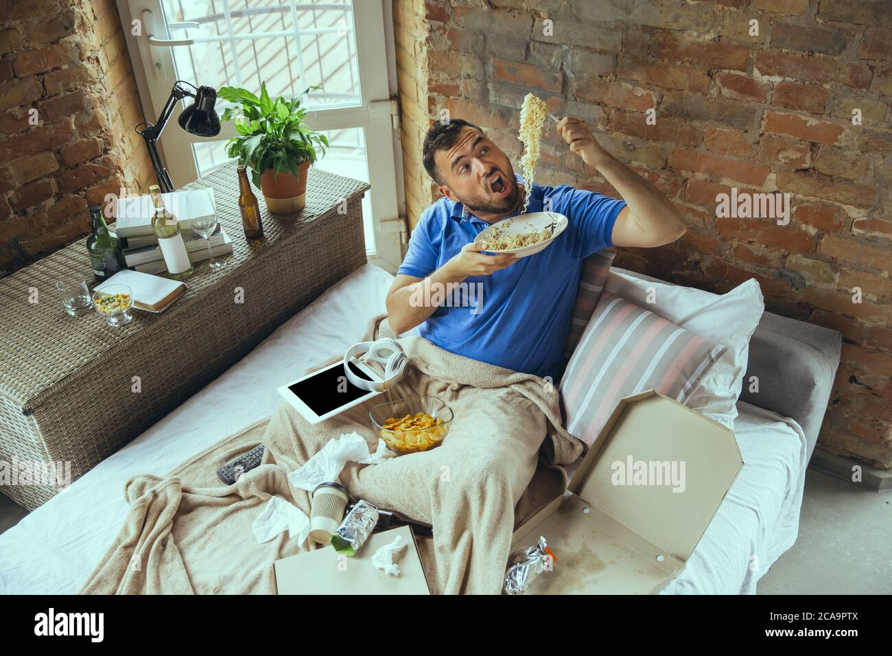 Eating instant noodles with funny hungry face. Lazy man living in his bed surrounded with messy. No need to go out to be happy. Using gadgets, watching movie and series, emotional. Fast food. Stock Photo