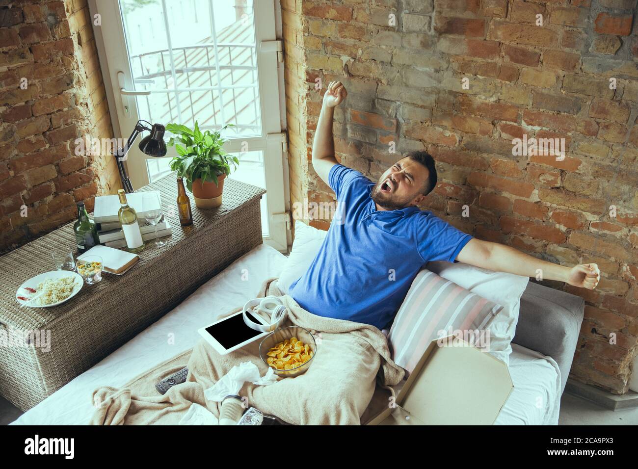 Yawns, stretches, feels full of happiness. Lazy man living in his bed surrounded with messy. No need to go out to be happy. Using gadgets, watching movie and series, emotional. Fast food. Stock Photo
