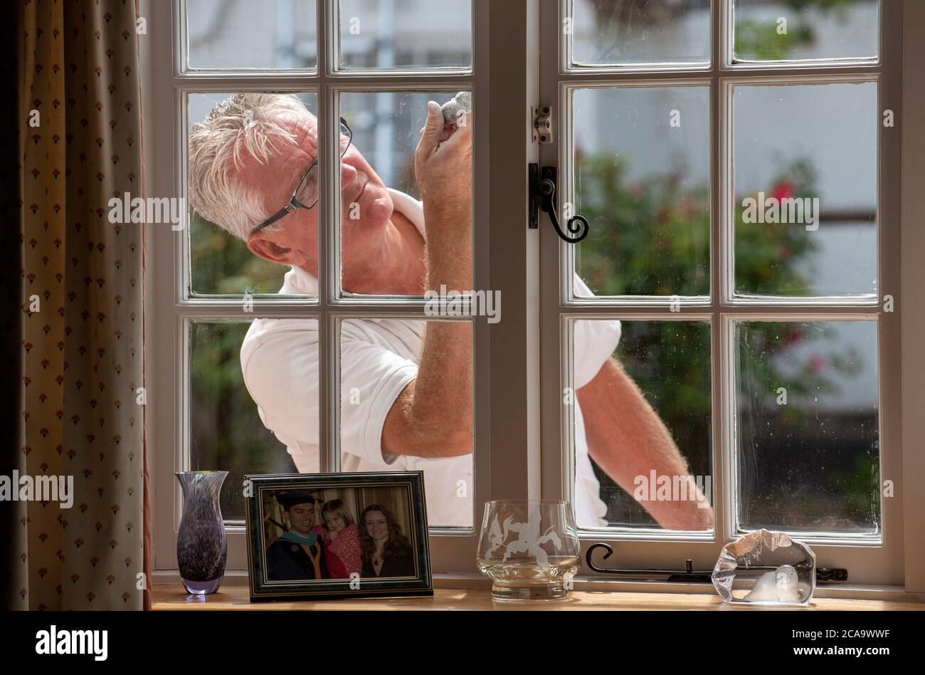 Hampshire, England, UK. 2020. Painter decorator painting small windows on a rural house viewed from inside the home. Stock Photo