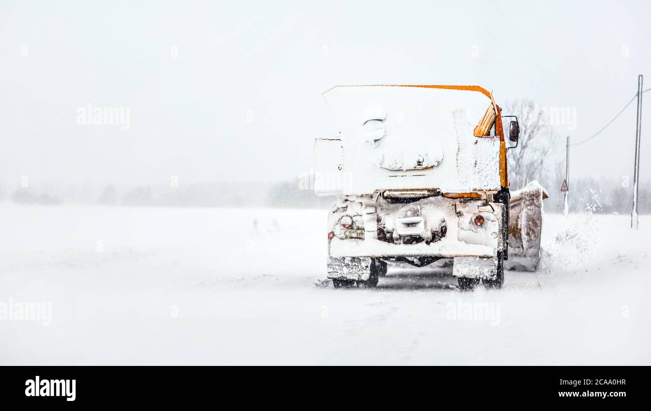 Orange plough truck on snow covered road, gray sky and trees in background, view from car driving behind Stock Photo