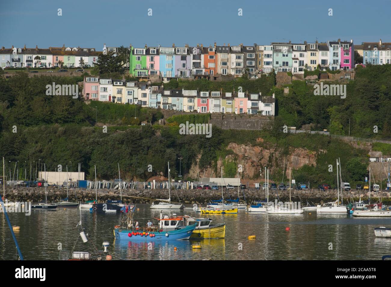Early AM Brixham harbour views. Row of colourful houses in two rows above harbour on skyline.  Boats on moorings below Blue sky Landscape format. Stock Photo