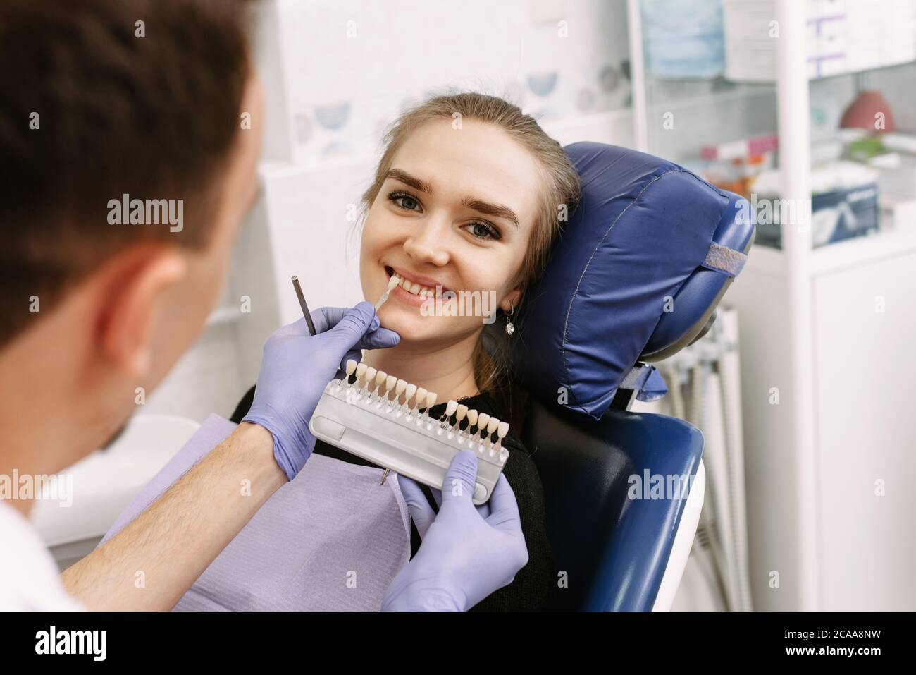 Dentist in blue medical gloves applying sample from tooth enamel scale to happy woman patient teeth to pick up right shade, teeth bleaching procedure, cropped, panorama. Stock Photo