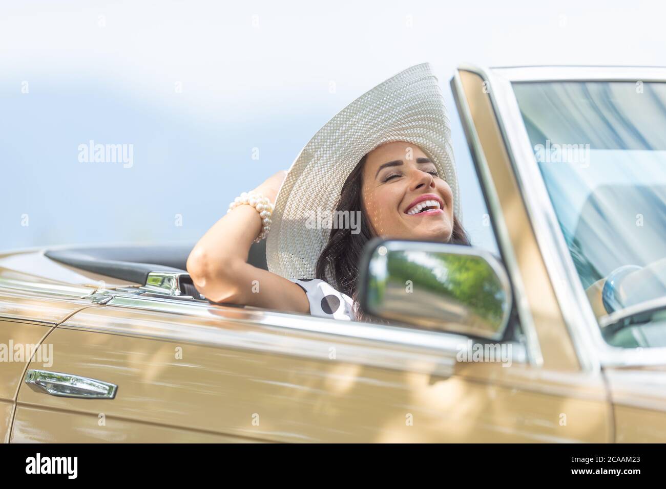 Right-handed female driver with eyes closed, head tilted backwards holds her hat and enjoys the summer cabiolet drive. Stock Photo