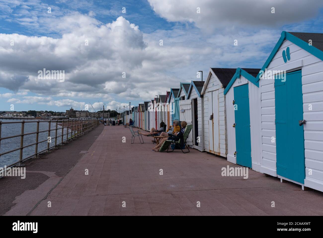 People sitting by Beach huts at Paignton enjoying the sun between the clouds on a typical British summer weather Stock Photo