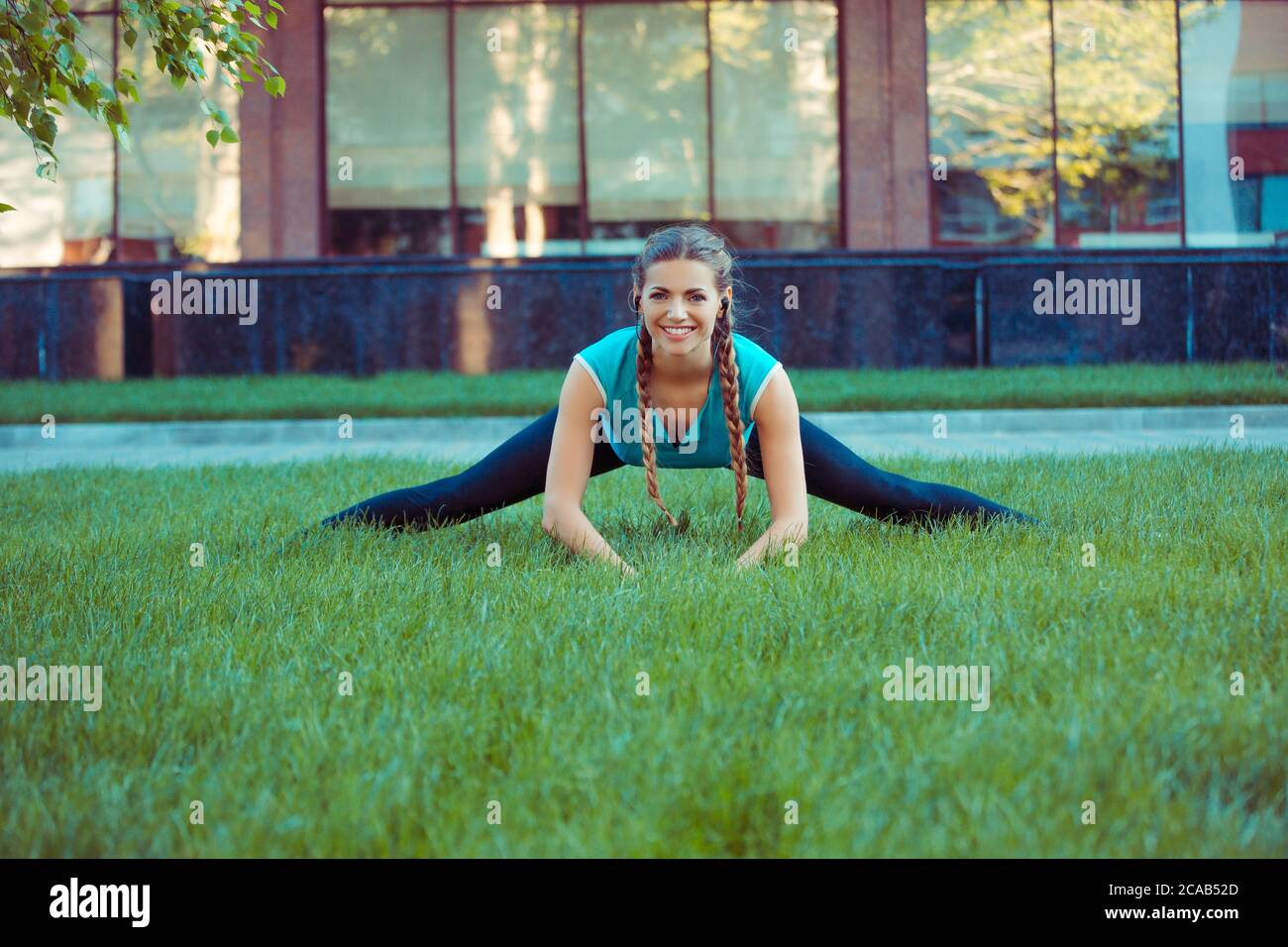 Pretty young brunette in sportswear stretching legs on green meadow standing in splits and smiling at camera Stock Photo
