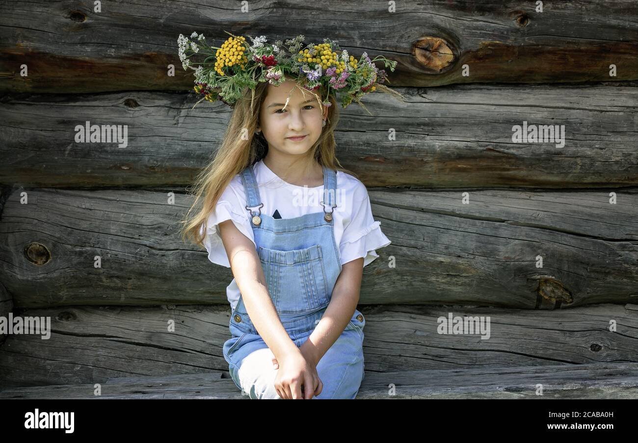 Summer portrait of a happy girl. Cute teenage girl in flower wreath. Background wall of logs. Stock Photo