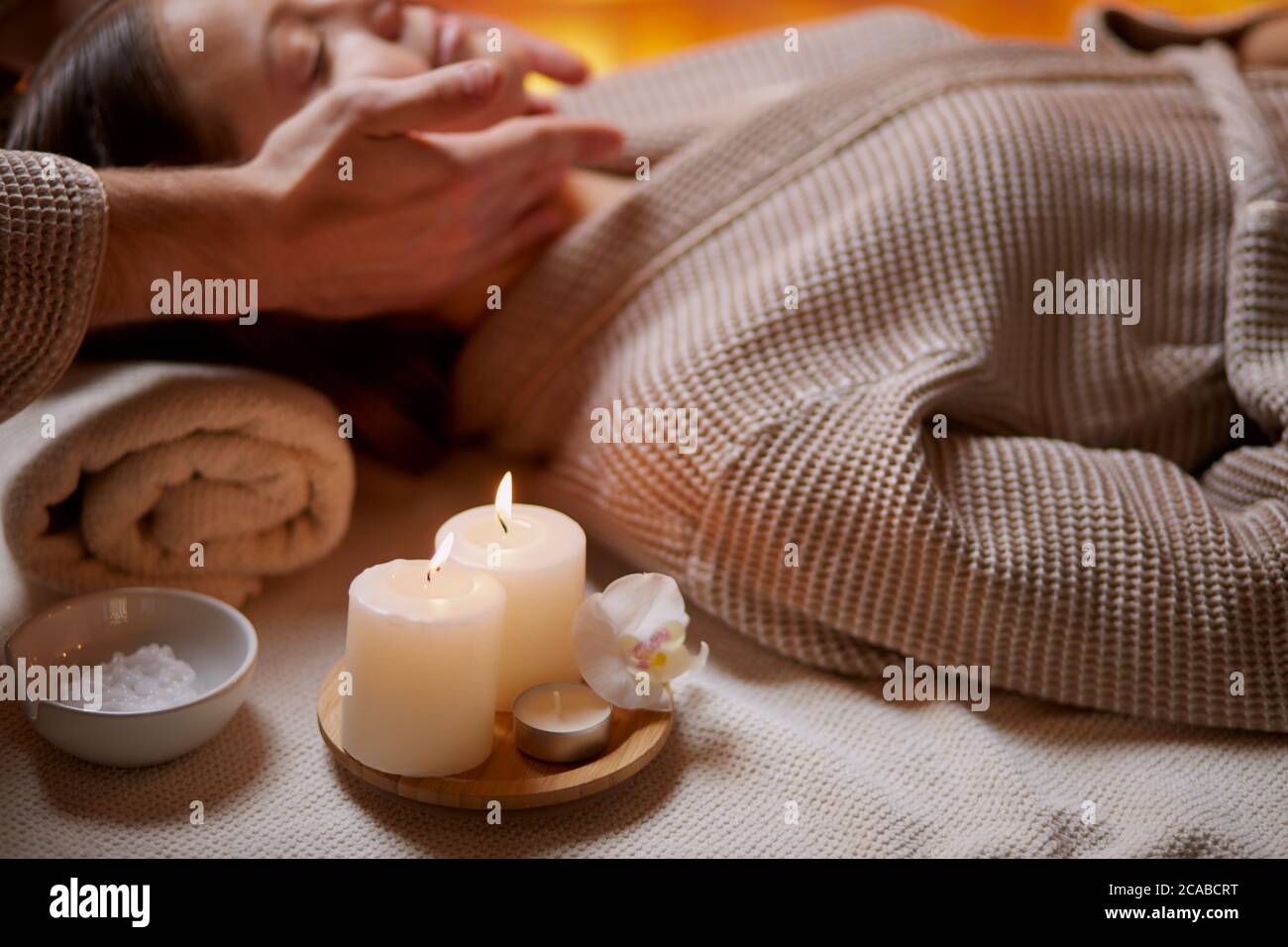 Young girl having massage on her face in spa salon. Wearing bathrobe, candles near her. Beauty treatment Stock Photo