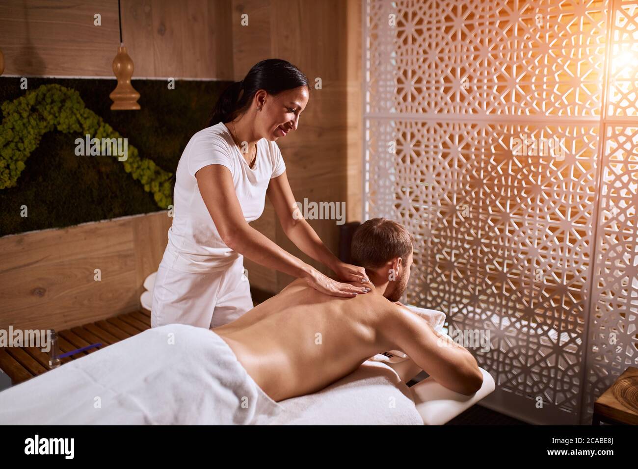 Charming pleasant woman wearing white uniform, working in spa hotel, massaging back of male client in brightly lighted room of hotel, total relaxation Stock Photo