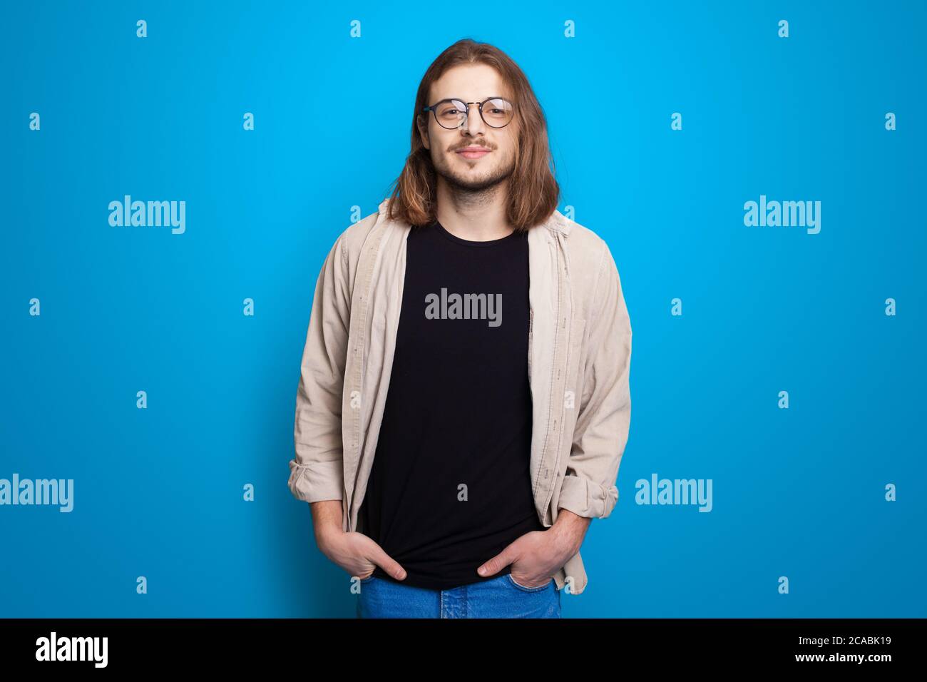 Fashionable man with long hair and bristle posing with eyeglasses on a blue studio wall Stock Photo