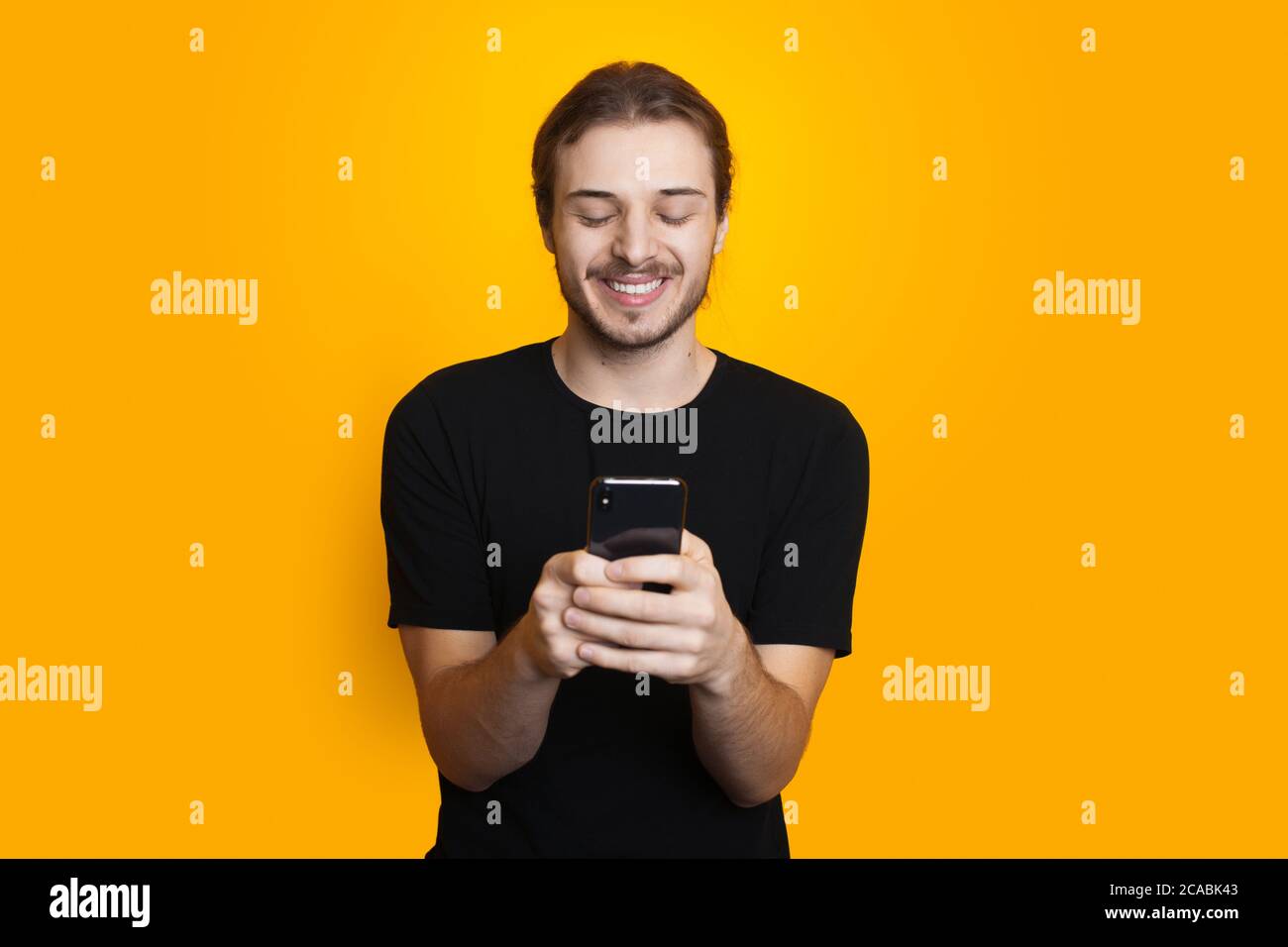 Bearded man with long hair is chatting on mobile in black clothes and smile on a yellow studio wall Stock Photo