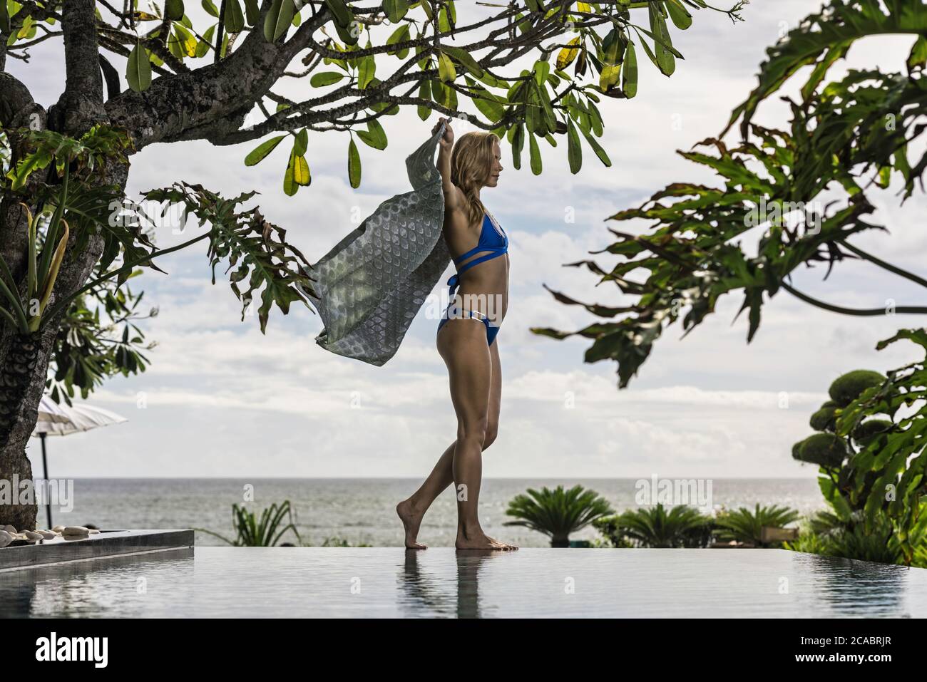 Asia, Indonesia, Bali, young beautiful Caucasian woman, wearing blue  bikini, walking along on edge of infinity pool, holding a sarong, with  tropical p Stock Photo - Alamy