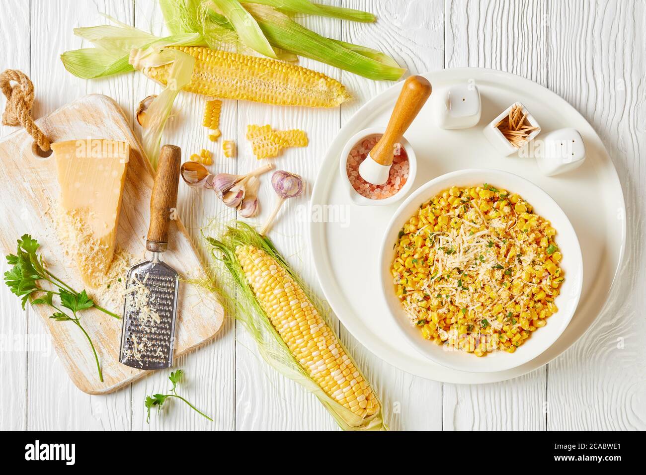 Parmesan Cilantro Corn in a white bowl on a white wooden table with ingredients on a white wooden table, horizontal view from above, flat lay Stock Photo