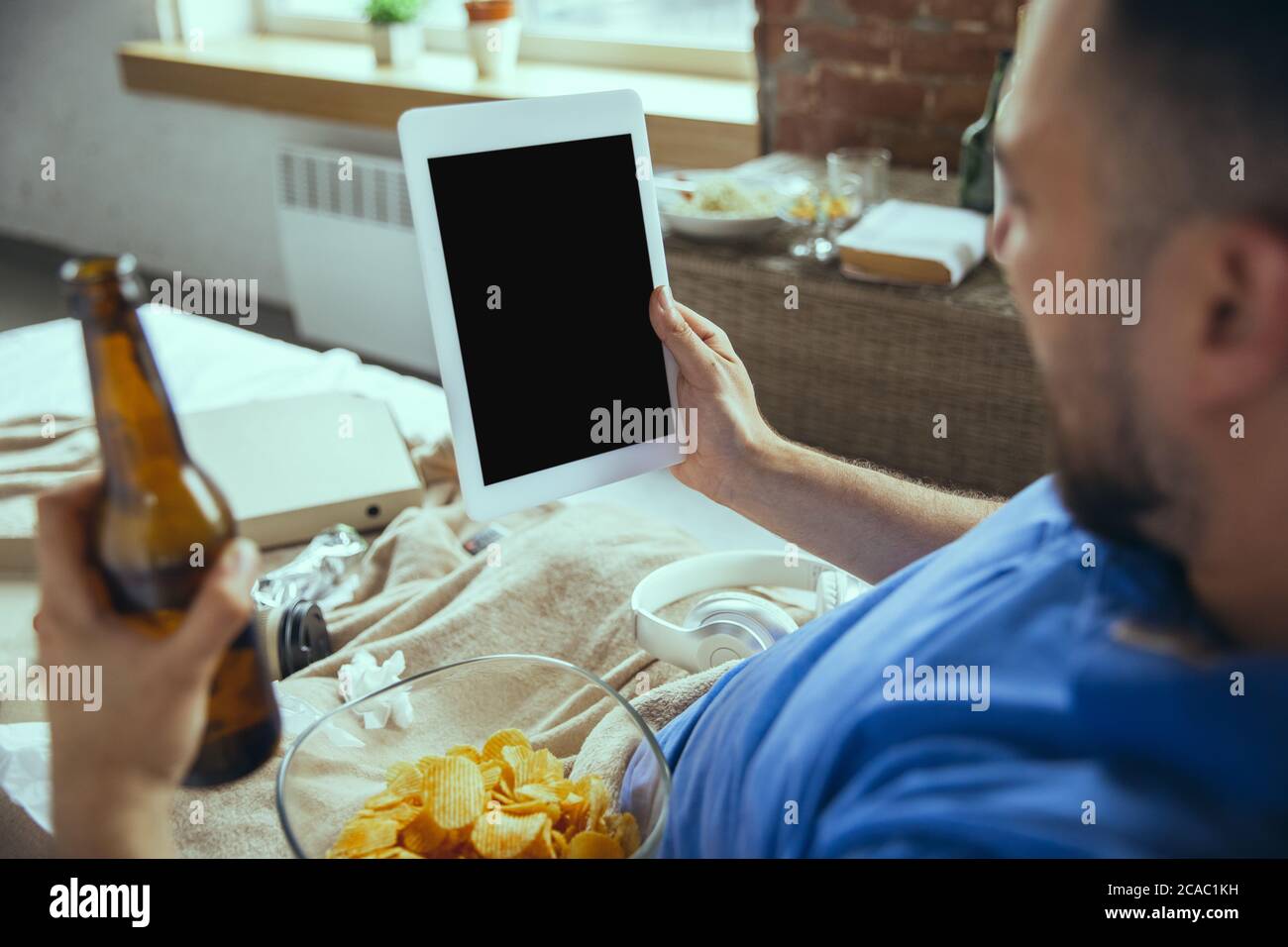 Sport fan, drinks beer. Lazy man living in his bed surrounded with messy. No need to go out to be happy. Using gadgets, watching movie and series, looks emotional. Tablet with copyspace for ad. Stock Photo