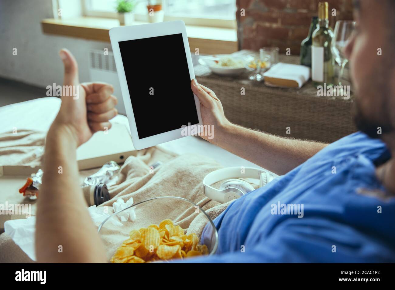 Eating crisps, thumb up. Lazy man living in his bed surrounded with messy. No need to go out to be happy. Using gadgets, watching movie and series, looks emotional. Tablet with copyspace for ad. Stock Photo