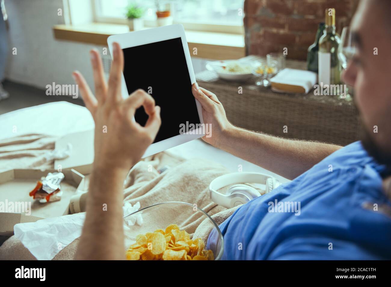 Eating crisps, nice gesture. Lazy man living in his bed surrounded with messy. No need to go out to be happy. Using gadgets, watching movie and series, looks emotional. Tablet with copyspace for ad. Stock Photo