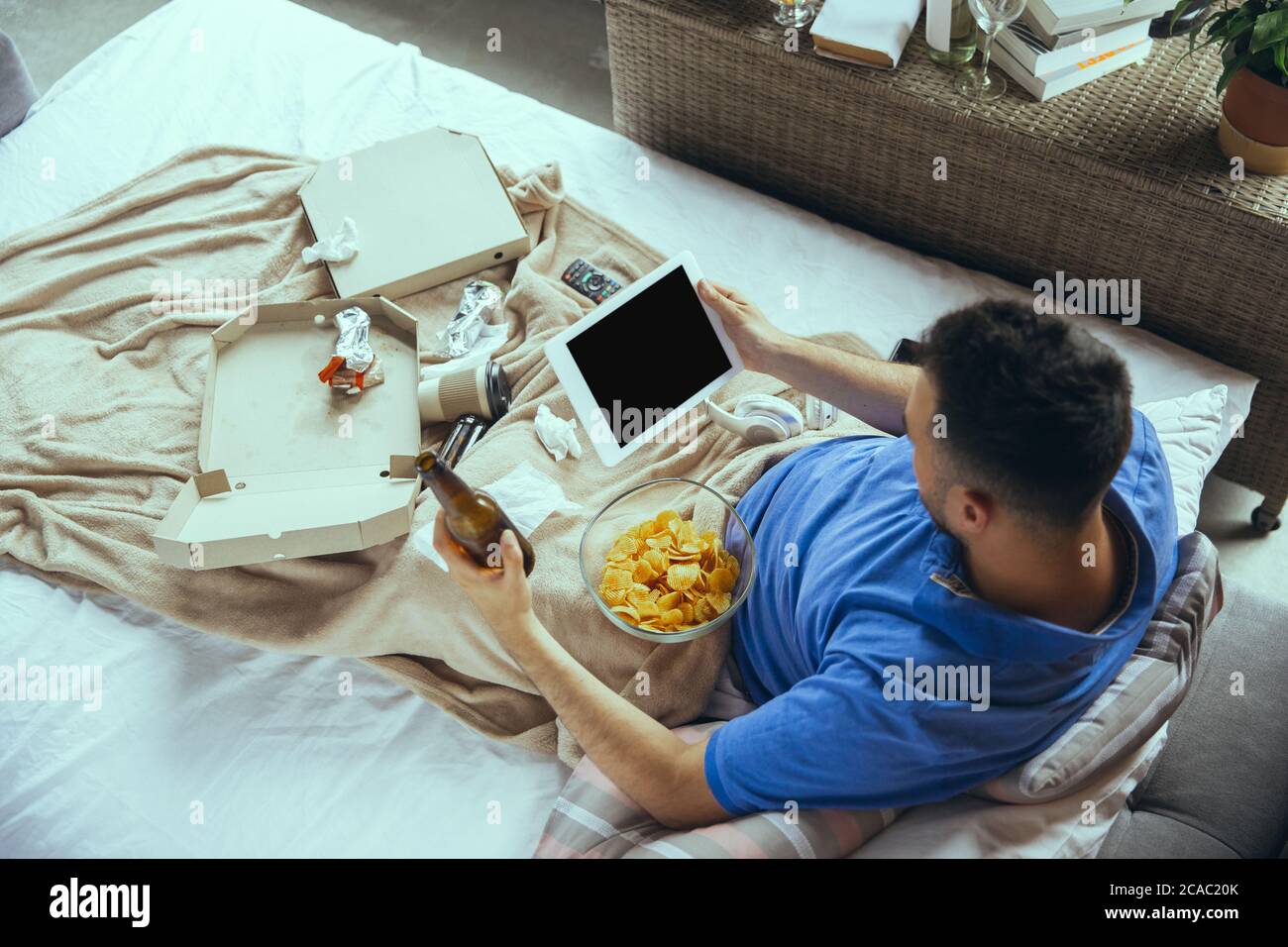 Eating crisps, drinks beer. Lazy man living in his bed surrounded with messy. No need to go out to be happy. Using gadgets, watching movie and series, looks emotional. Tablet with copyspace for ad. Stock Photo