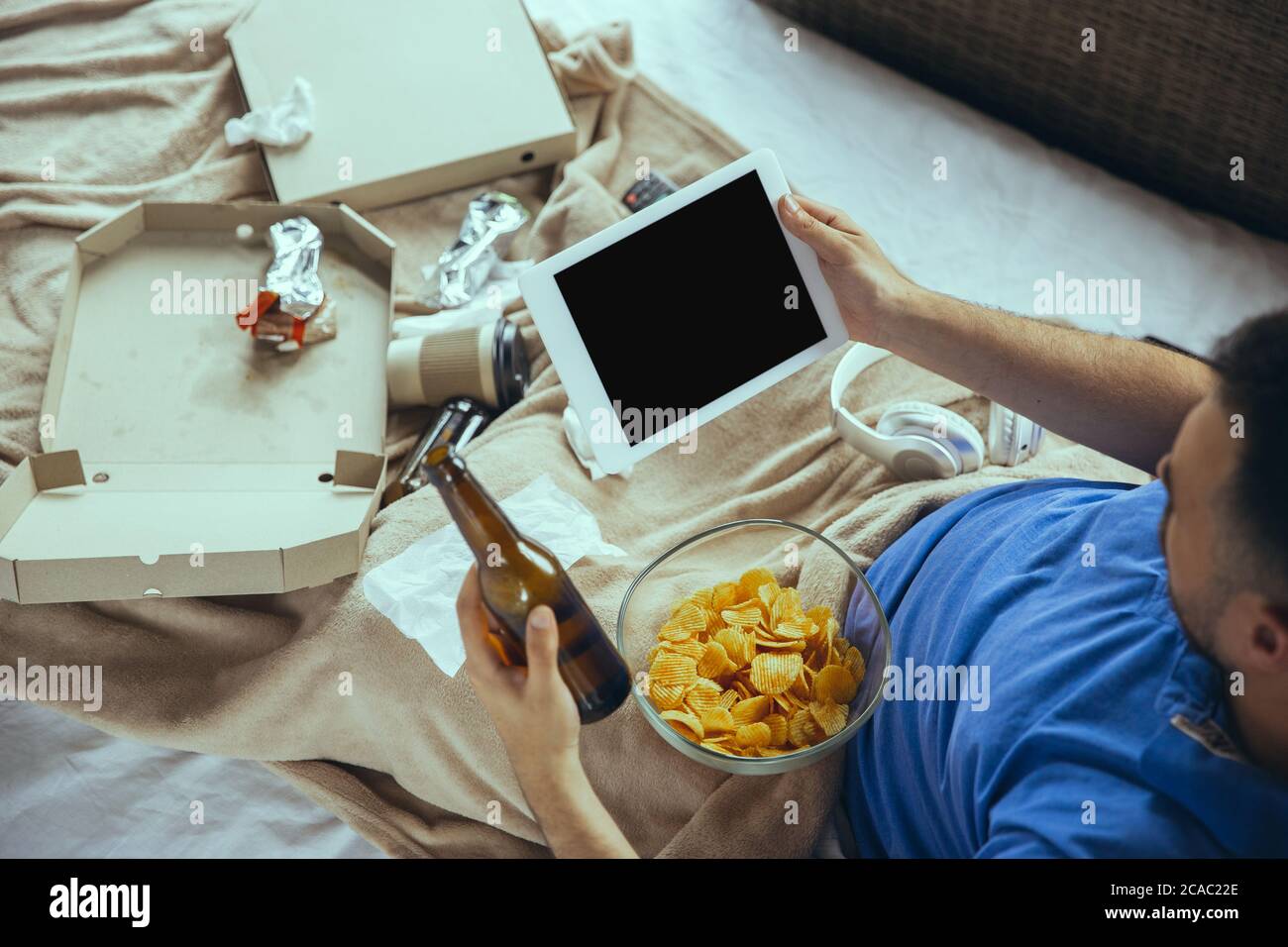 Eating crisps, drinks beer. Lazy man living in his bed surrounded with messy. No need to go out to be happy. Using gadgets, watching movie and series, looks emotional. Tablet with copyspace for ad. Stock Photo