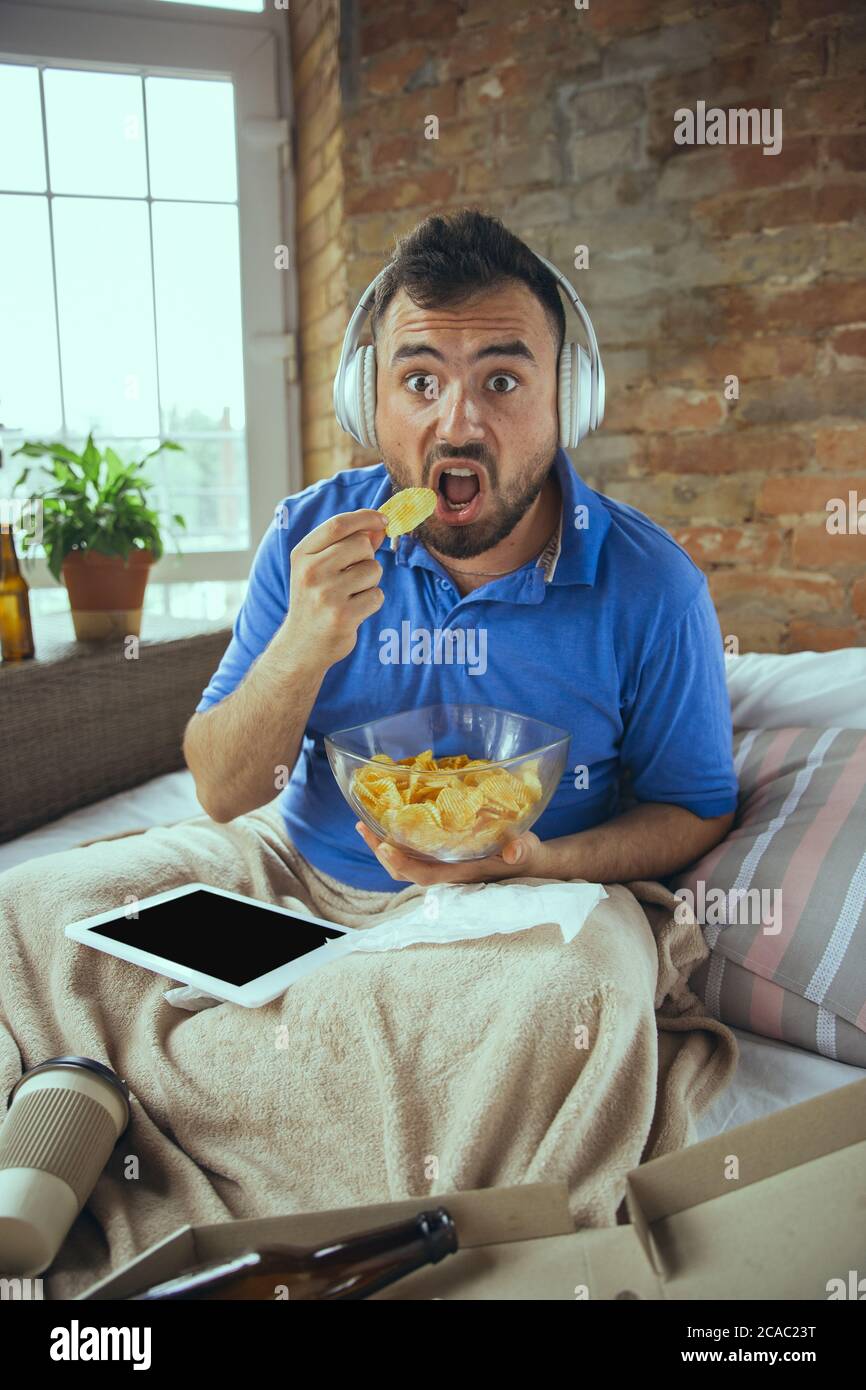 Eating crisps, shocked. Lazy man living in his bed surrounded with messy. No need to go out to be happy. Using gadgets, watching movie and series, looks emotional. Tablet with copyspace for ad. Stock Photo