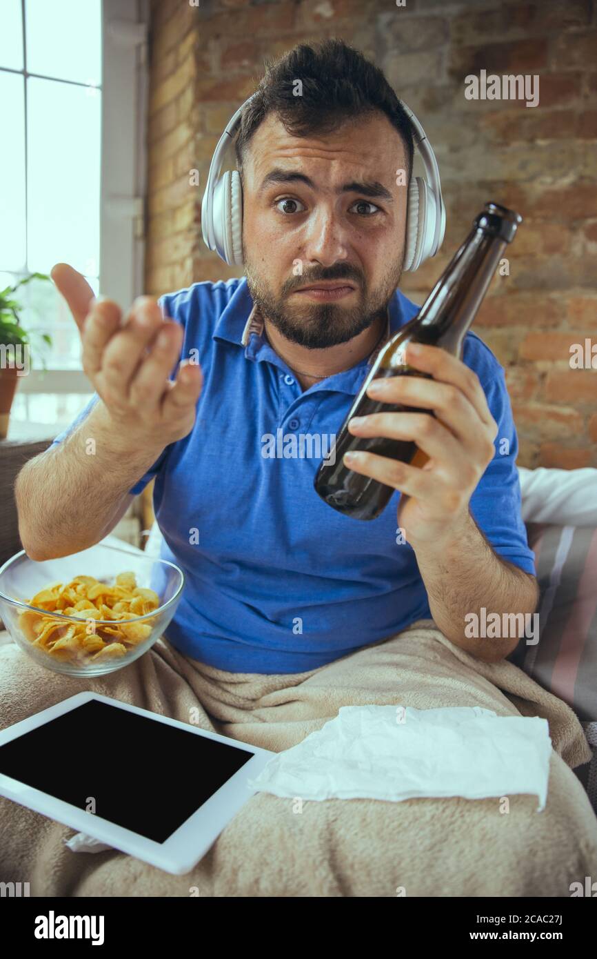 Asking, drinks beer. Lazy man living in his bed surrounded with messy. No need to go out to be happy. Using gadgets, watching movie and series, looks emotional. Tablet with copyspace for ad. Stock Photo