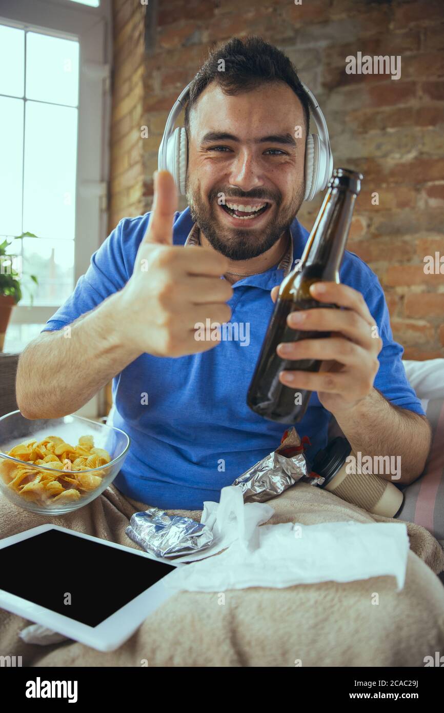 Thumb up, drinks beer. Lazy man living in his bed surrounded with messy. No need to go out to be happy. Using gadgets, watching movie and series, looks emotional. Tablet with copyspace for ad. Stock Photo
