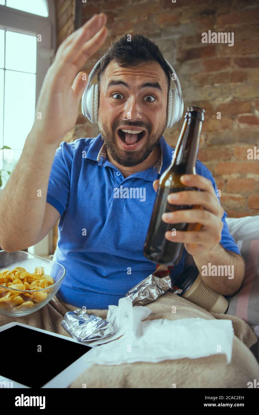 Cheering, happy, drinks beer. Lazy man living in his bed surrounded with messy. No need to go out to be happy. Using gadgets, watching movie and series, looks emotional. Tablet with copyspace for ad. Stock Photo