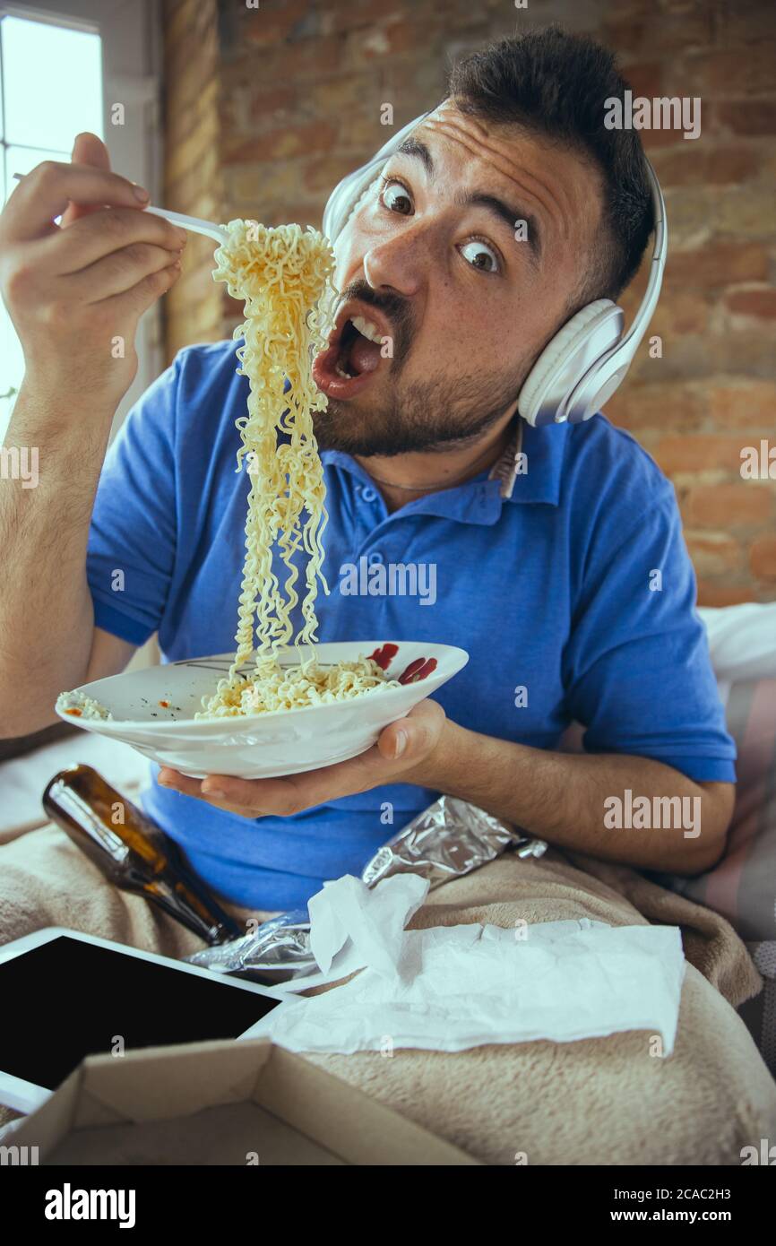 Hungry eating instant noodles. Lazy man living in his bed surrounded with messy. No need to go out to be happy. Using gadgets, watching movie and series, looks emotional. Tablet with copyspace for ad. Stock Photo