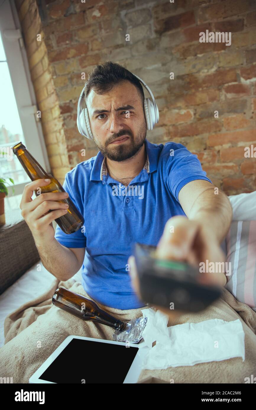 Bored using TV remote, drinks beer. Lazy man living in his bed surrounded with messy. No need to go out to be happy. Using gadgets, watching movie and series, looks emotional. Tablet with copyspace for ad. Stock Photo