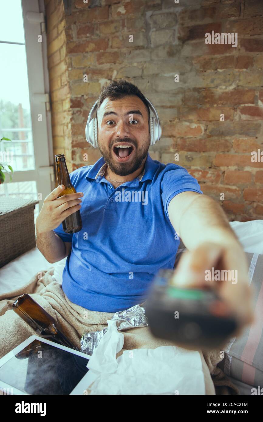 Happy using TV remote, drinks beer. Lazy man living in his bed surrounded with messy. No need to go out to be happy. Using gadgets, watching movie and series, looks emotional. Tablet with copyspace for ad. Stock Photo