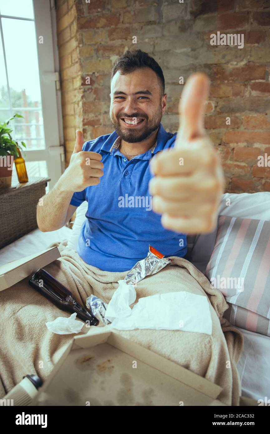 Smiling, thumbs up. Lazy caucasian man living in his bed surrounded with messy. No need to go out to be happy. Using gadgets, watching movie and series, social media, looks emotional. Home lifestyle. Stock Photo