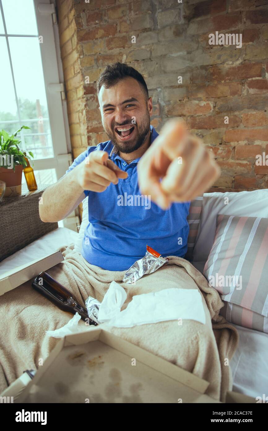 Pointing, exciting. Lazy caucasian man living in his bed surrounded with messy. No need to go out to be happy. Using gadgets, watching movie and series, social media, looks emotional. Home lifestyle. Stock Photo