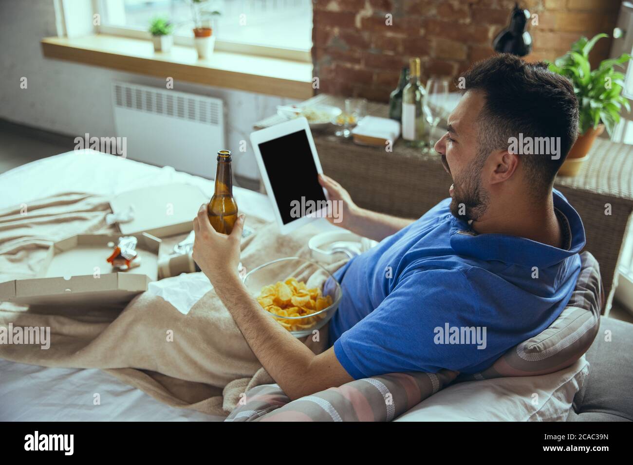 Cheering, screaming. Lazy caucasian man living in his bed surrounded with messy. No need to go out to be happy. Using gadgets, watching movie and series, social media, looks emotional. Home lifestyle. Stock Photo
