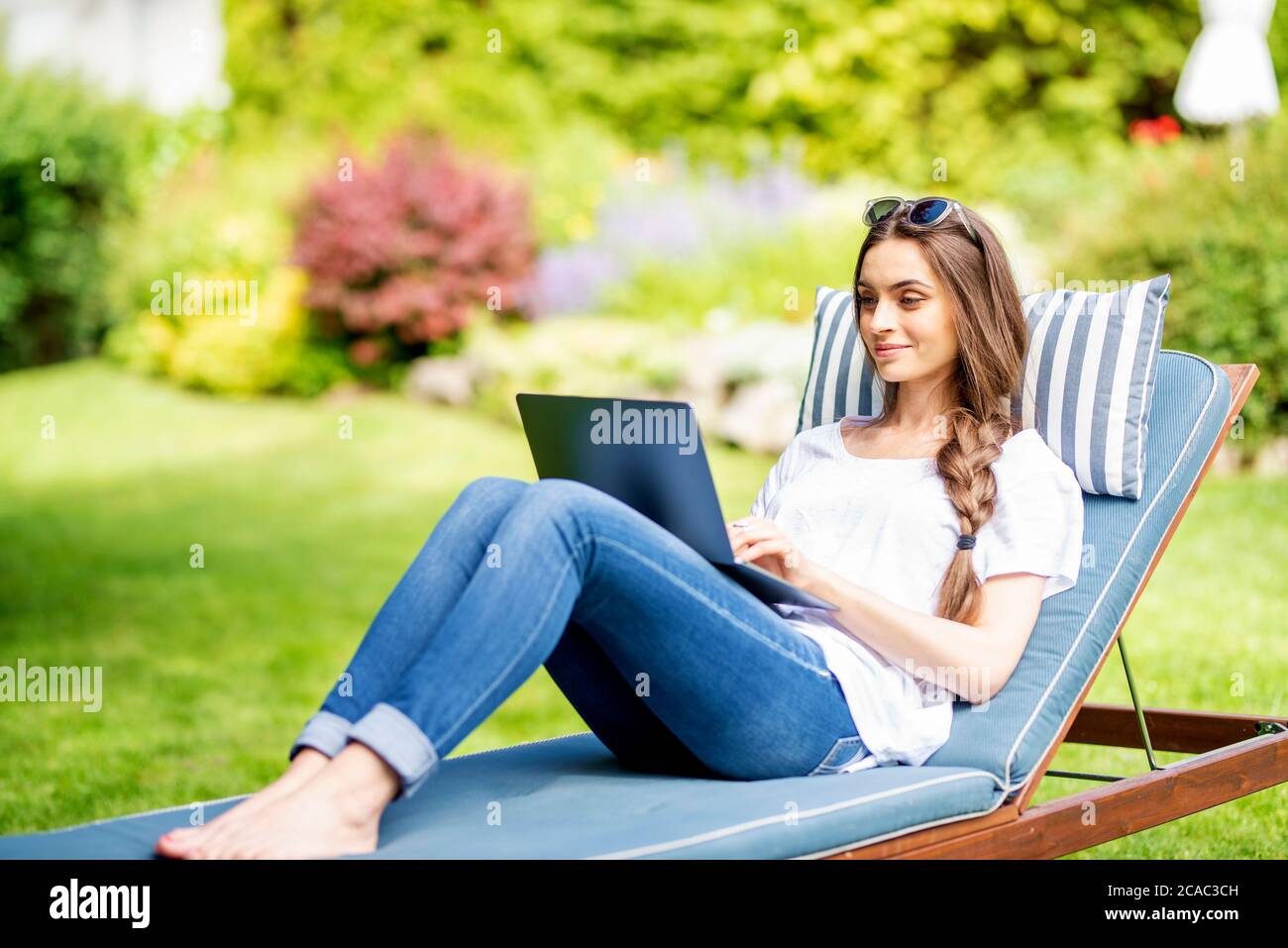 Shot of happy young woman sitting on sunbed in the garden and using her ...