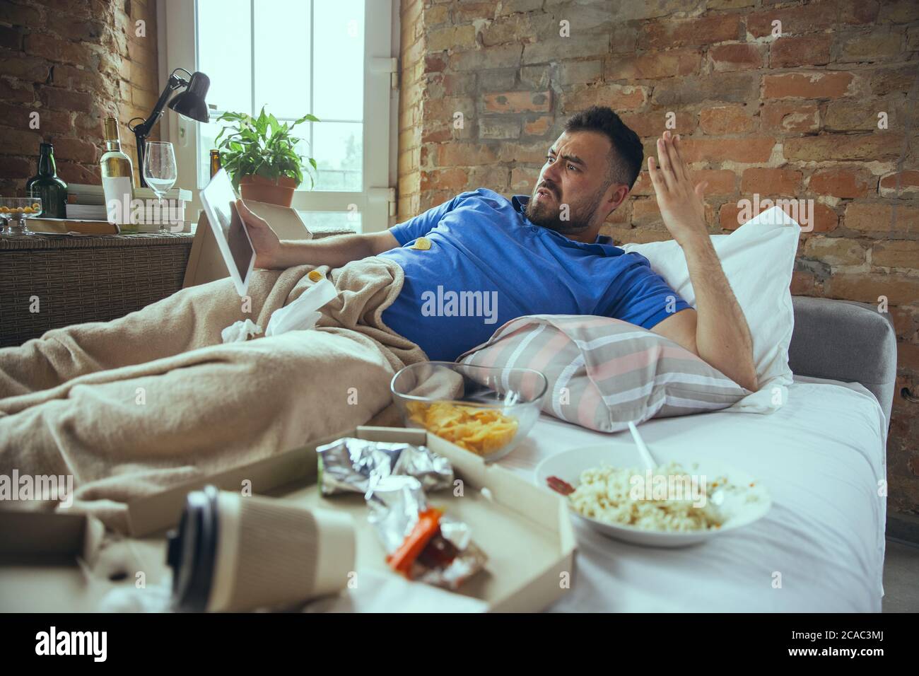 Angry watching tablet. Lazy caucasian man living in his bed surrounded with messy. No need to go out to be happy. Using gadgets, watching movie and series, social media, looks emotional. Home lifestyle. Stock Photo