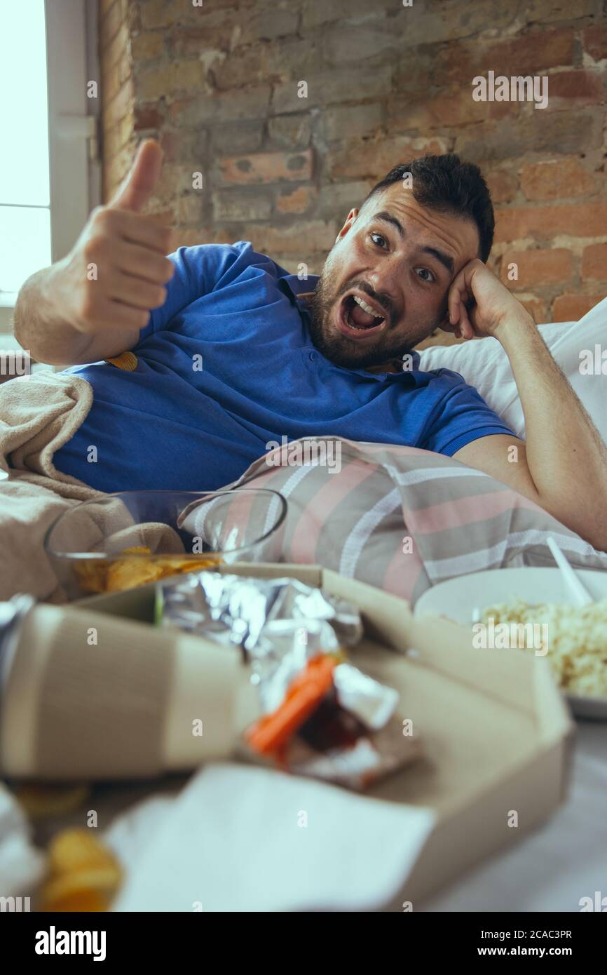 Thumb up, nice, laughting. Lazy caucasian man living in his bed surrounded with messy. No need to go out to be happy. Using gadgets, watching movie and series, social media, looks emotional. Home lifestyle. Stock Photo