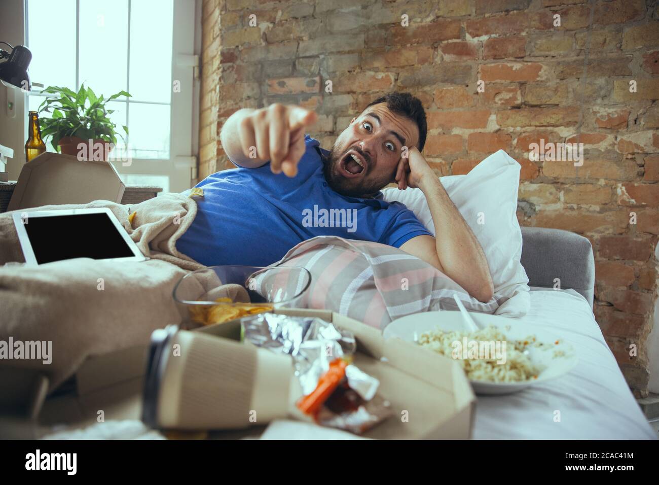 Pointing, laughting. Lazy caucasian man living in his bed surrounded with messy. No need to go out to be happy. Using gadgets, watching movie and series, social media, looks emotional. Home lifestyle. Stock Photo