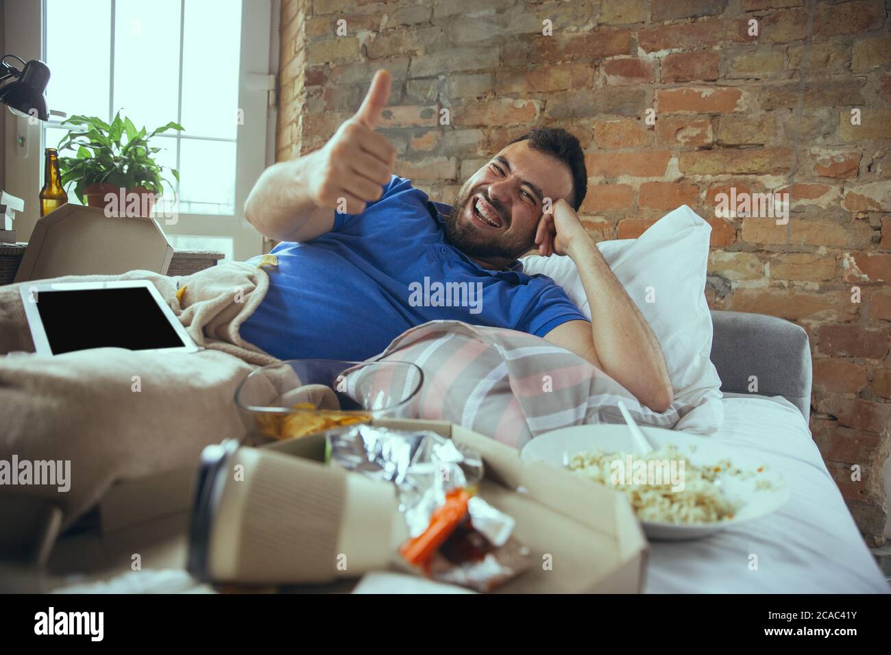 Thumb up, laughting. Lazy caucasian man living in his bed surrounded with messy. No need to go out to be happy. Using gadgets, watching movie and series, social media, looks emotional. Home lifestyle. Stock Photo