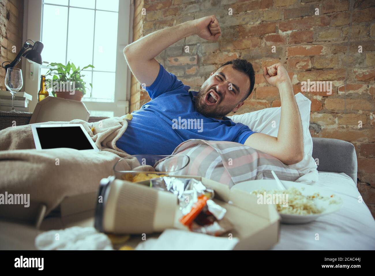 Celebrating, winner. Lazy caucasian man living in his bed surrounded with messy. No need to go out to be happy. Using gadgets, watching movie and series, social media, looks emotional. Home lifestyle. Stock Photo