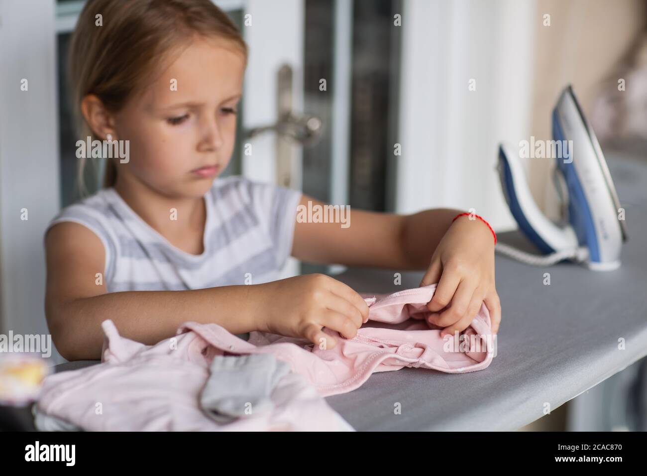 Cute little girl with blonde hair is leaning on ironing clothes on